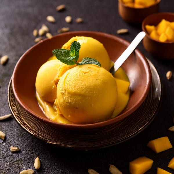 Three scoops of golden mango ice cream with mint in a brown bowl, surrounded by cardamom pods.