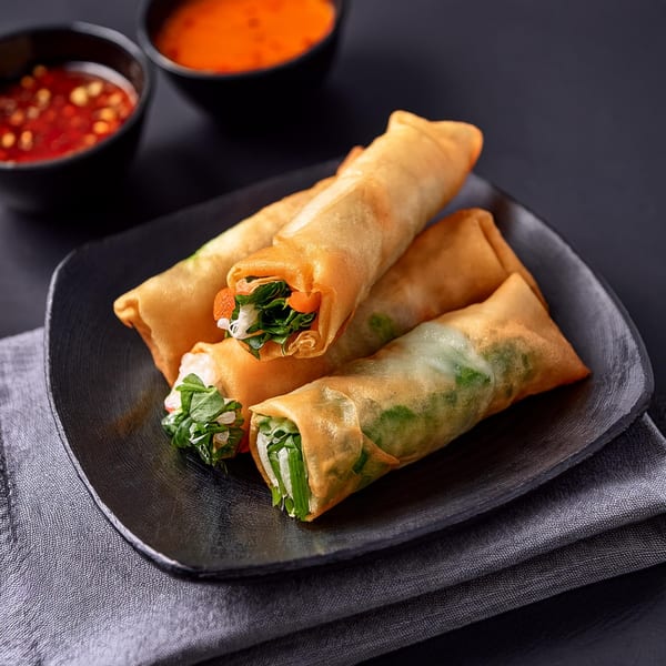 Four fresh spring rolls with green filling and two bowls of dipping sauce on a dark plate.